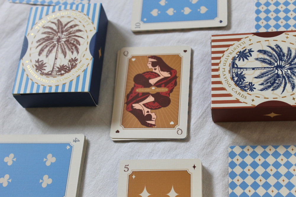 A styled flat-lay of a card game deck and cards on a white textured fabric background. The deck box is navy blue with a cream palm tree emblem and blue-and-white stripes.