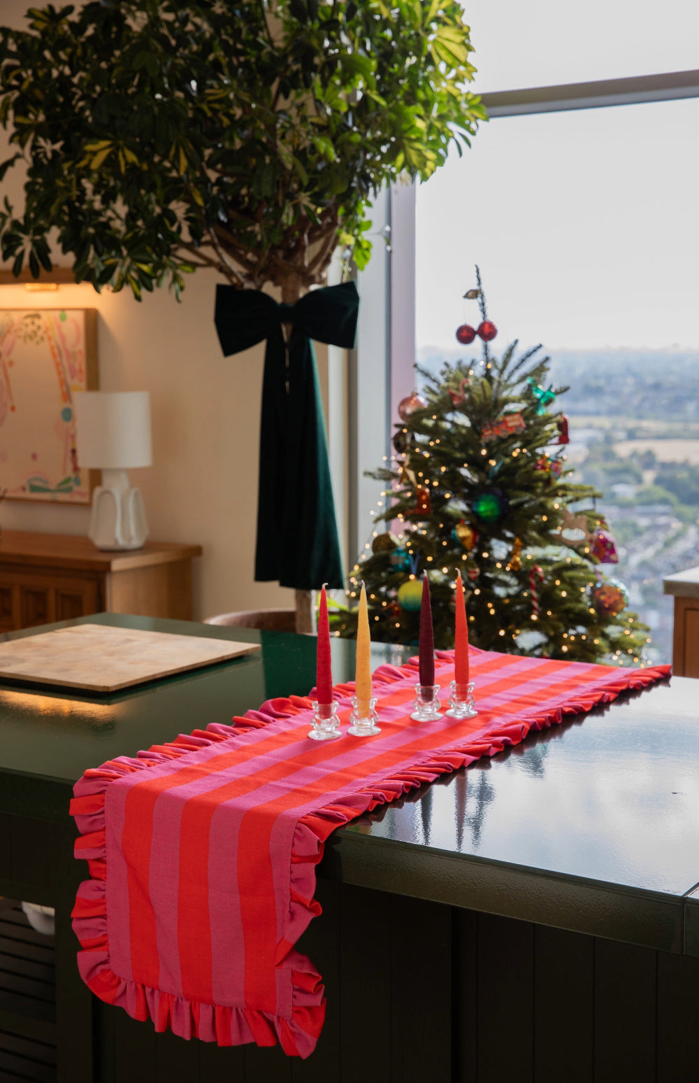 A bright pink and red striped table runner with ruffled edges draped over a dark table in a decorated holiday room, with candles, a Christmas tree, and a window view in the background.