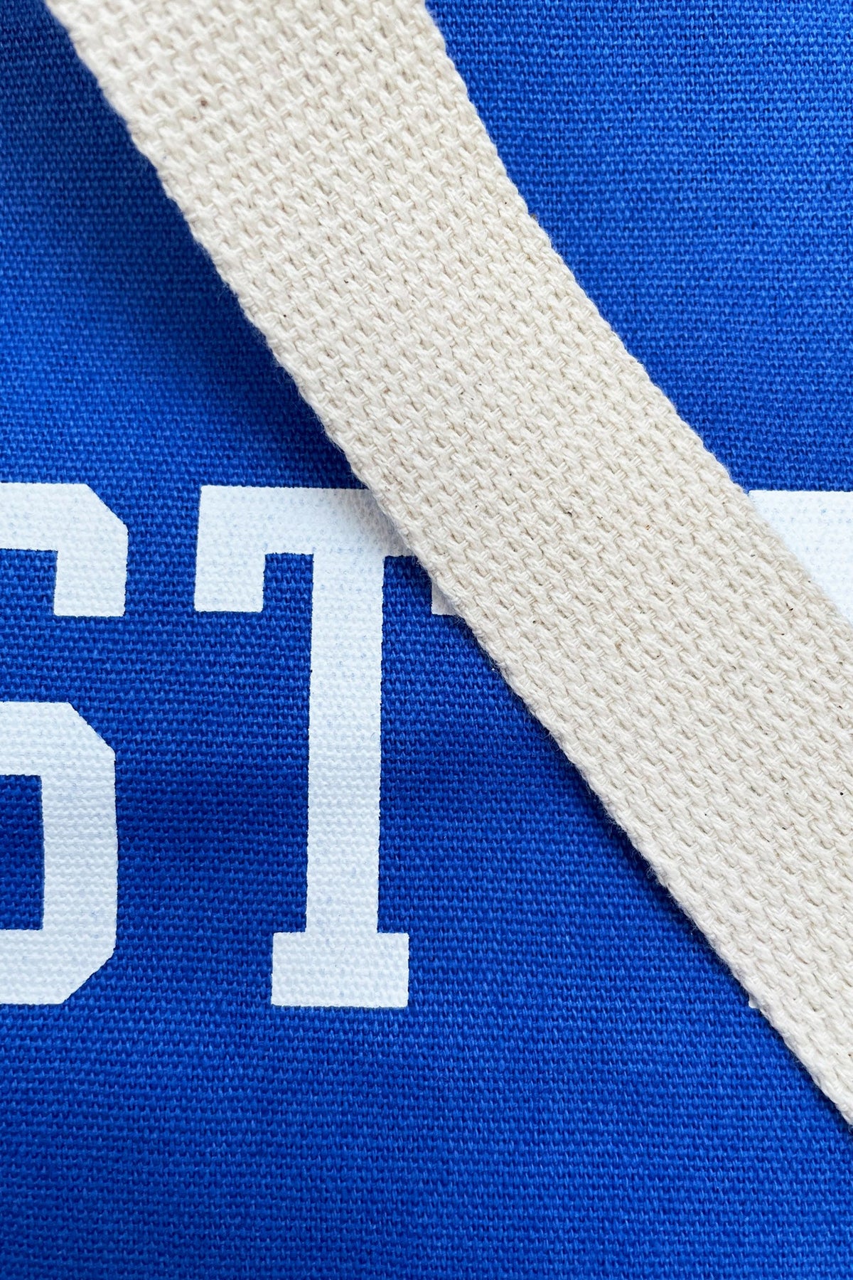 Close-up detail of a blue woven fabric item with a thick off-white canvas strap diagonally crossing the frame and partial white lettering visible.