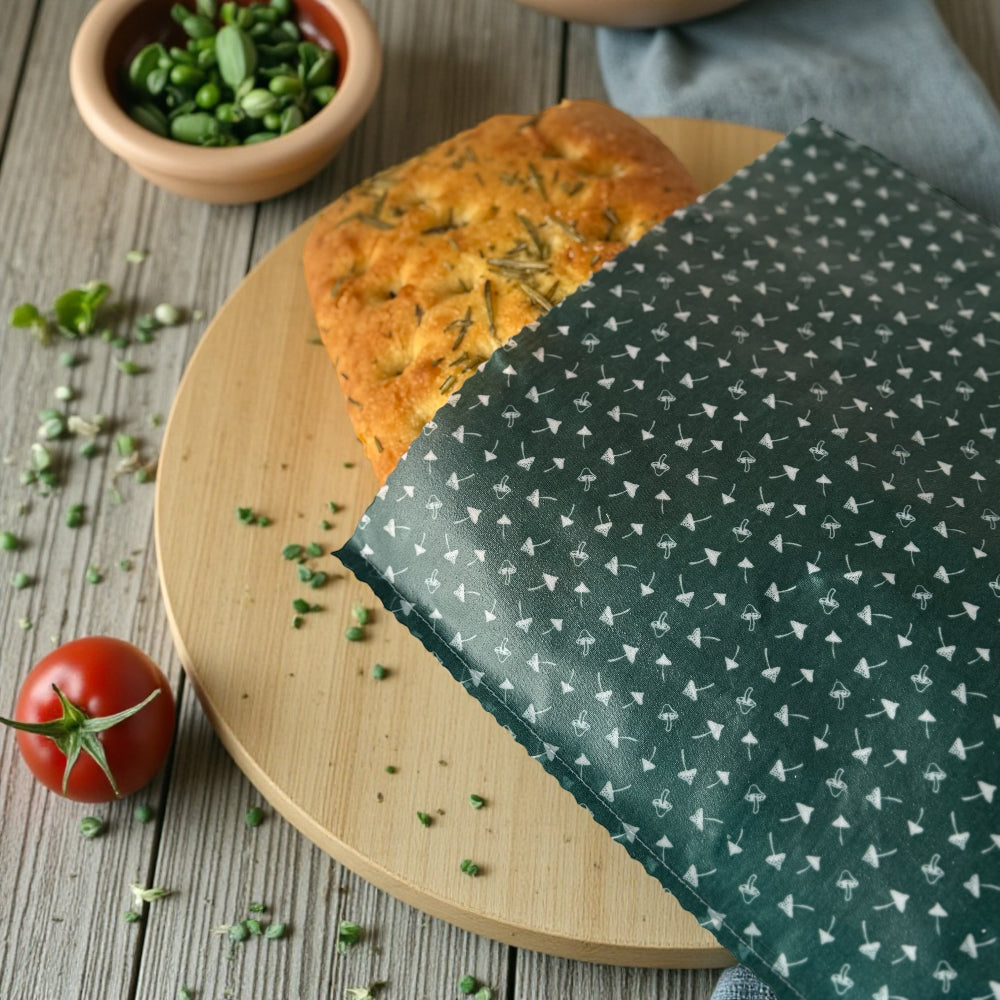 Loaf of bread on a wooden board with a green patterned toadstool beeswax wrap, surrounded by fresh herbs and a tomato.