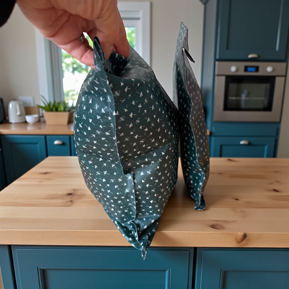 Two beeswax food storage bags on a wooden kitchen counter with a person's hand holding one of them.