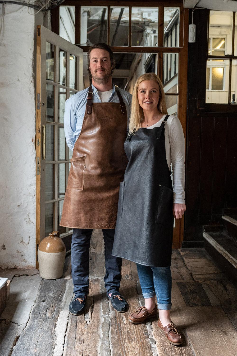 Two people wearing aprons standing in a rustic interior setting.