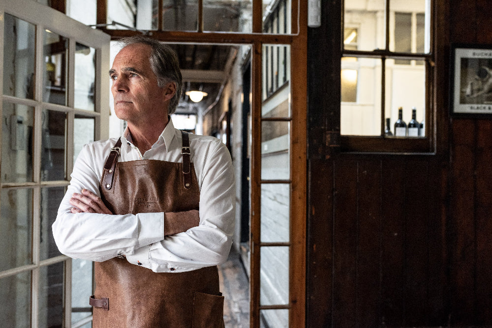 Man wearing a brown apron standing in a room with large windows.