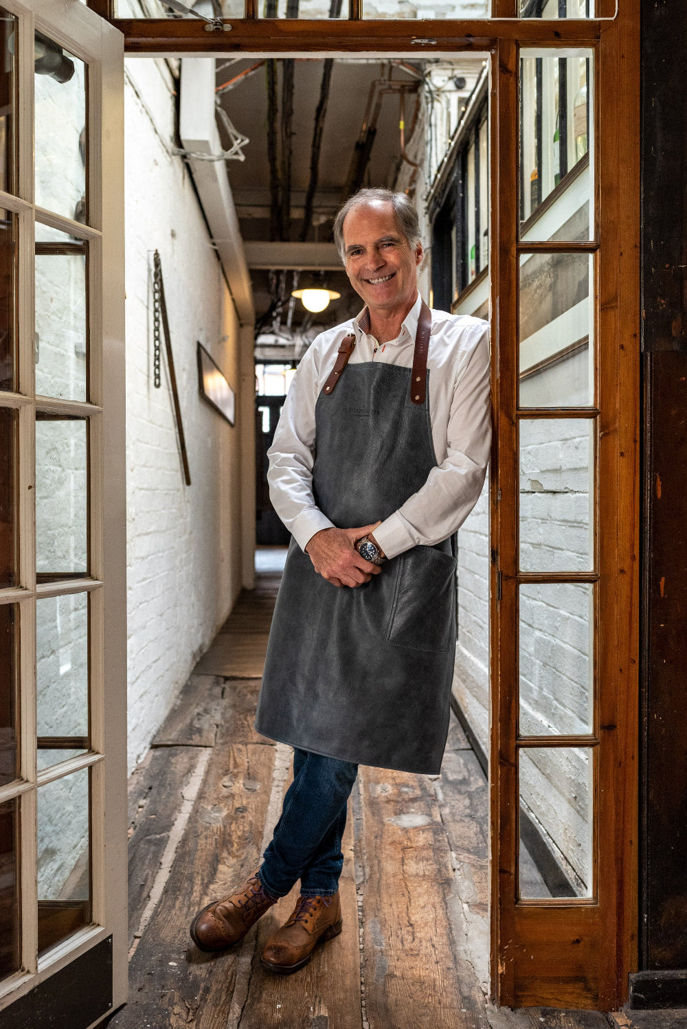 Man wearing a grey apron standing in a doorway with wooden floors and white walls.