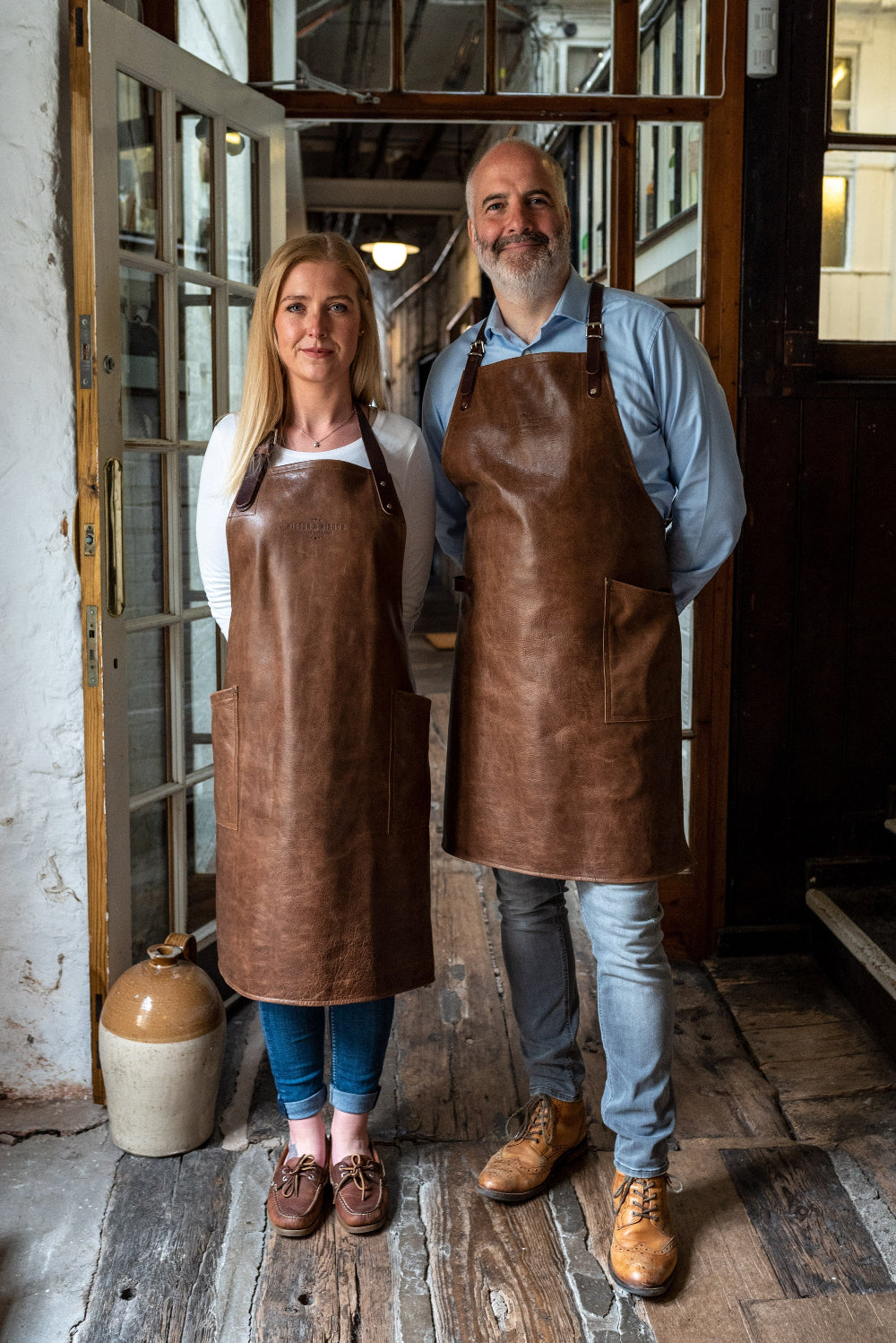 Two people wearing brown aprons standing in a rustic interior setting.