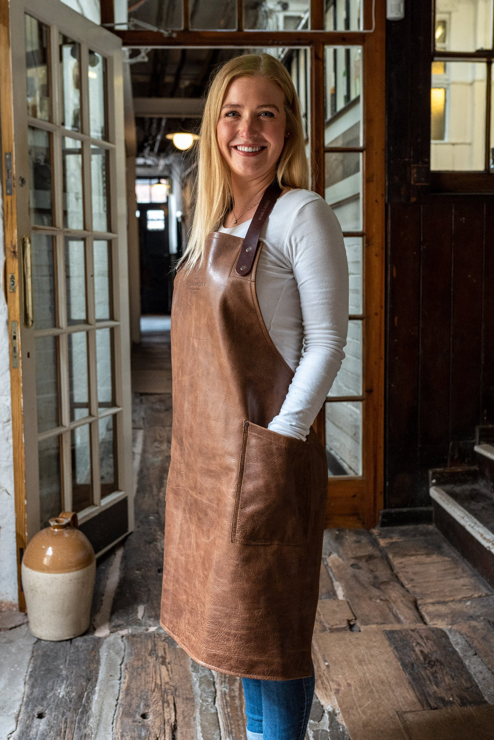 Woman wearing a brown leather apron standing in a rustic interior setting.