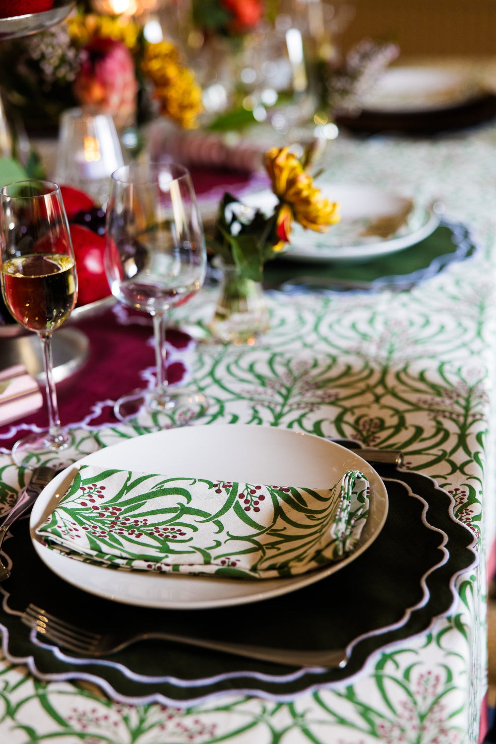 A styled table setting featuring a white tablecloth with a green botanical damask pattern, layered with dark green and black charger plates, a white plate, and a matching patterned napkin. Wine glasses and blurred flowers are visible in the background.