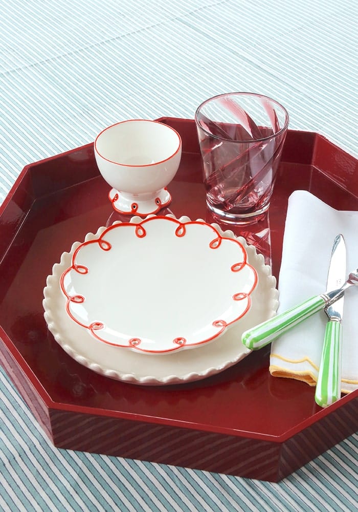 Red tray with white ceramic scalloped edge plate, glass, and cutlery on a striped background
