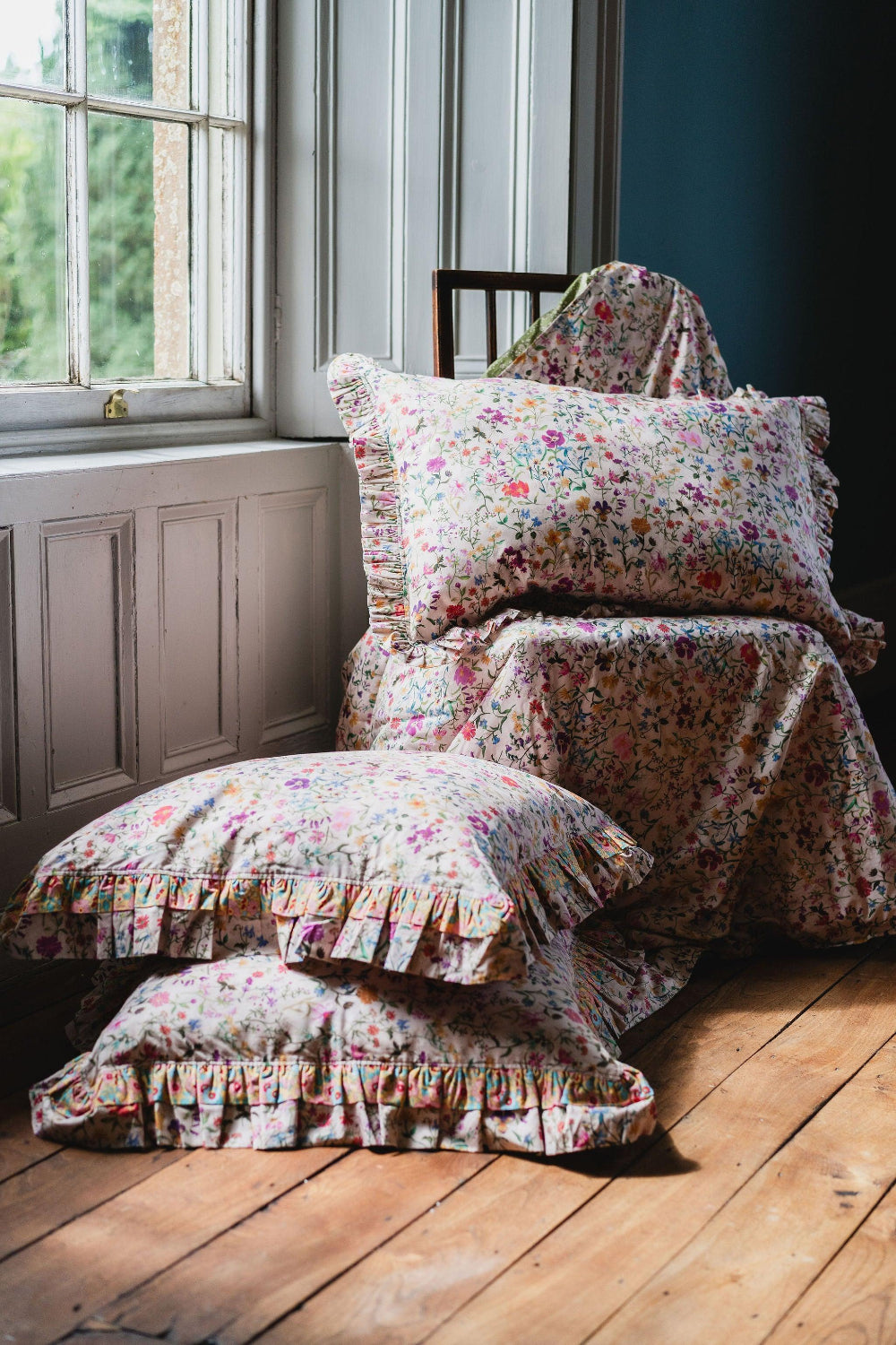 Floral patterned cushions and blanket on a chair by a window with natural light.