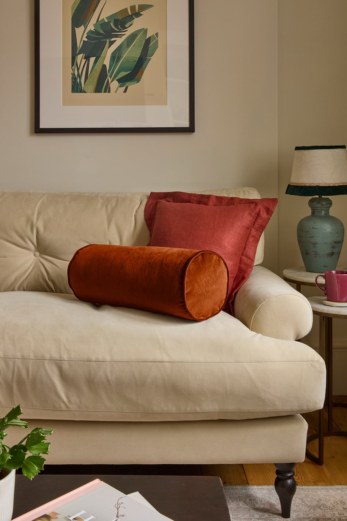 A styled living room scene featuring a cream-colored sofa with two decorative pillows: a cylindrical rust-orange velvet pillow in front of a rust-red pillow. A framed botanical print hangs on the beige wall above, and a blue-green table lamp sits on a side table to the right.