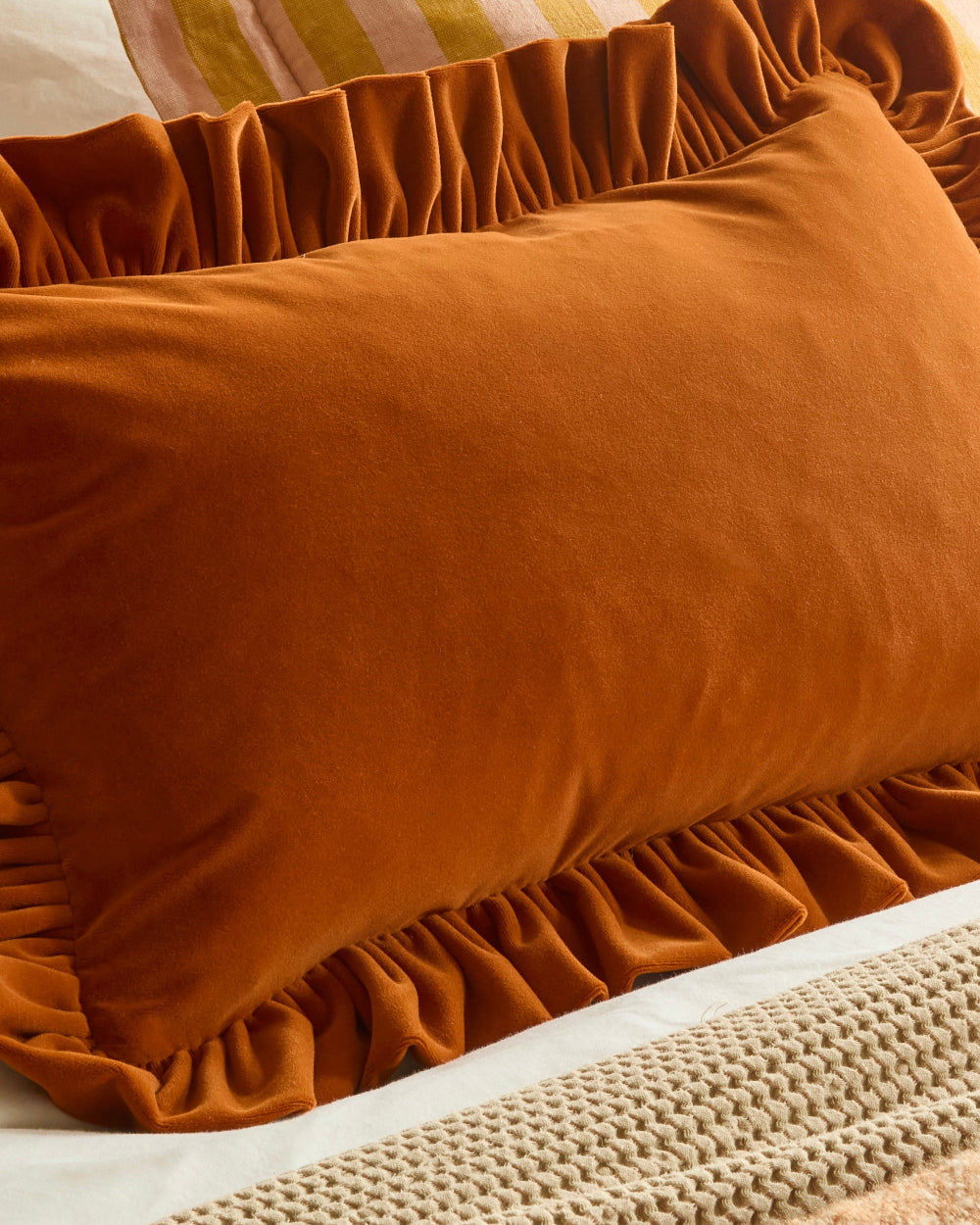 A close-up of a rust-orange velvet pillow with ruffled trim, resting on a bed with a beige knit blanket and striped pillows in the background.