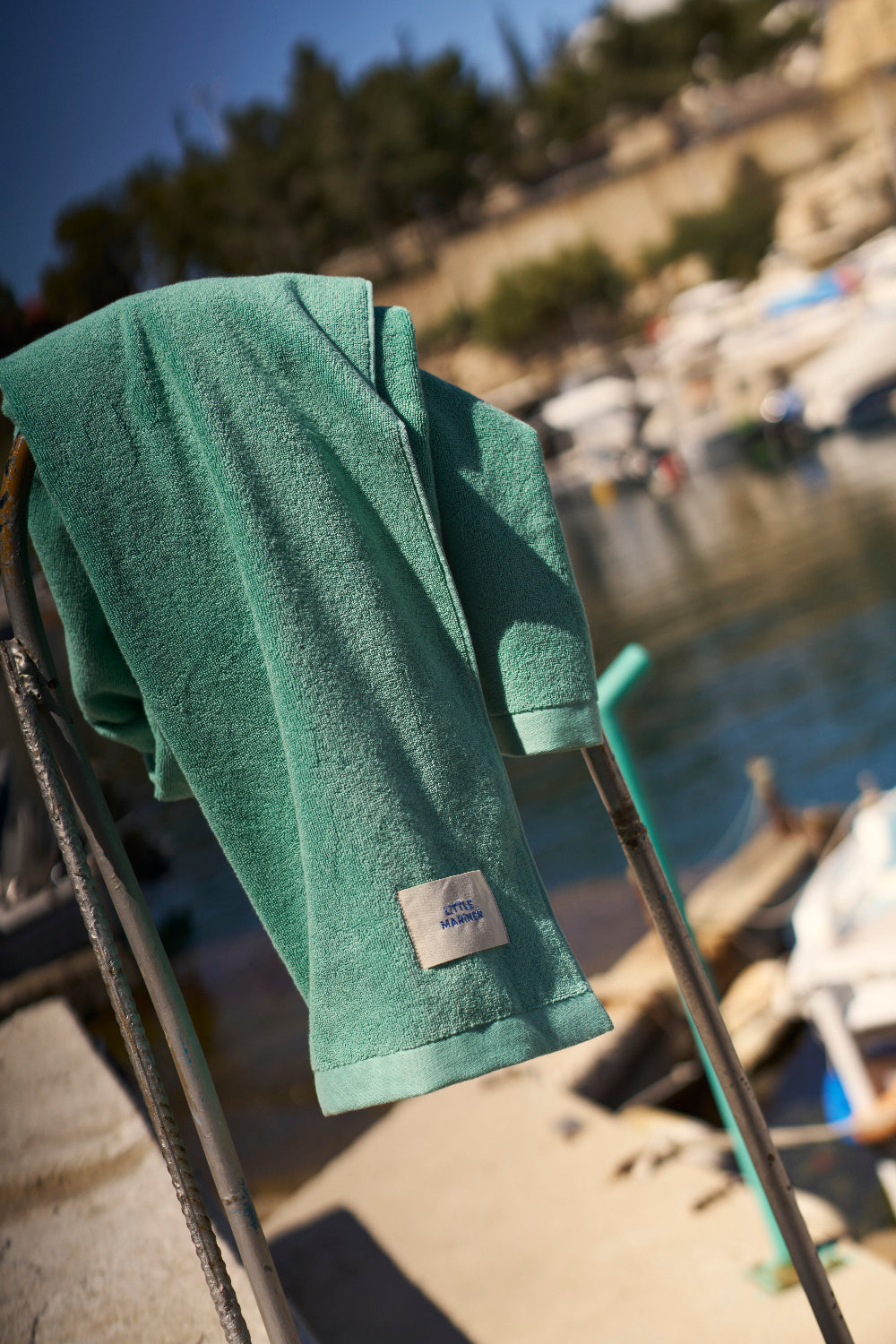 Green towel with a brand label on a chair by a waterfront with boats.