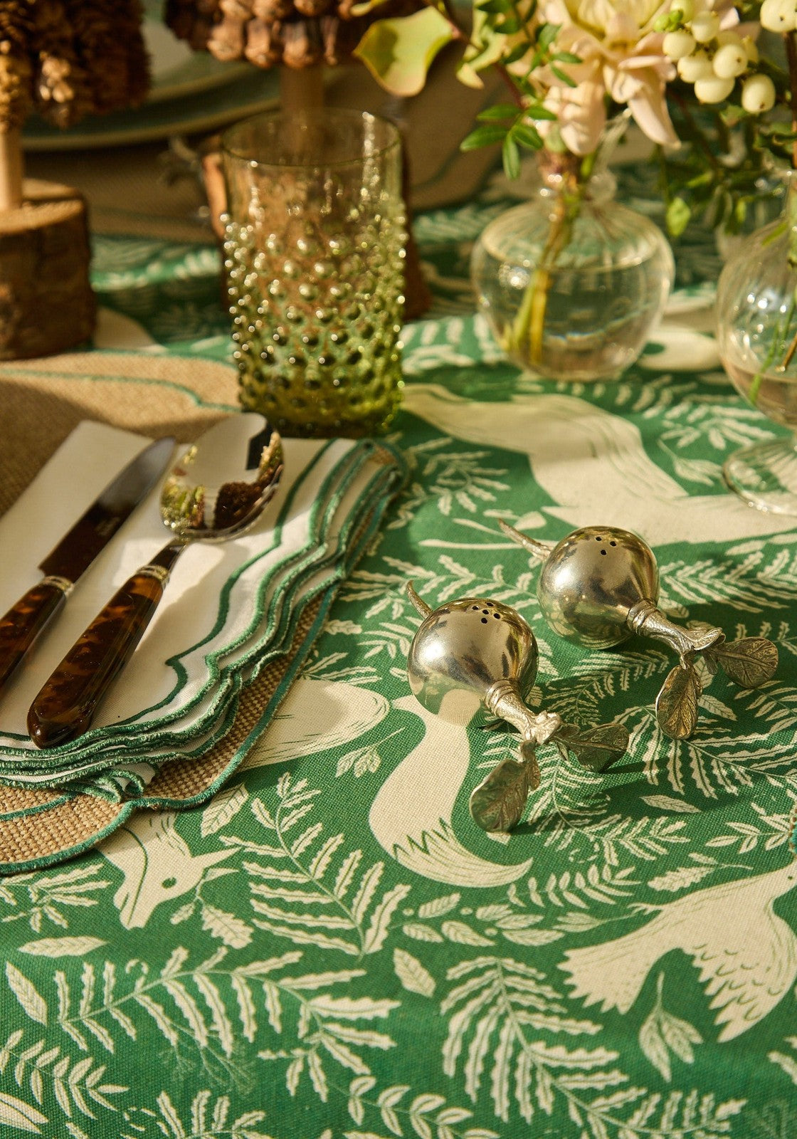 Table setting with a green and white patterned tablecloth, silver salt and pepper shakers, and floral arrangements.