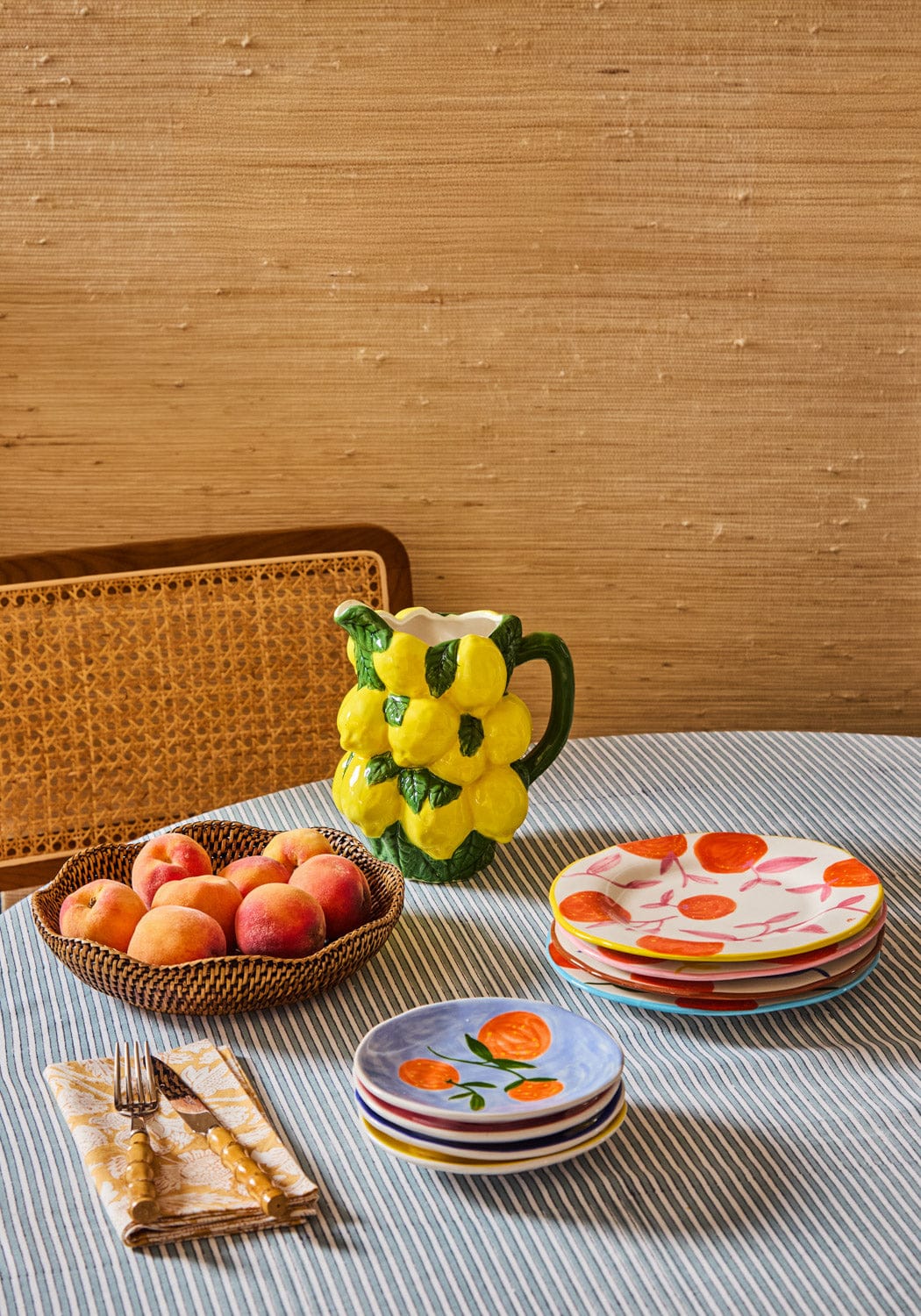 Table setting with fruit-themed plates, a pitcher, and a basket of apples against a wooden wall.