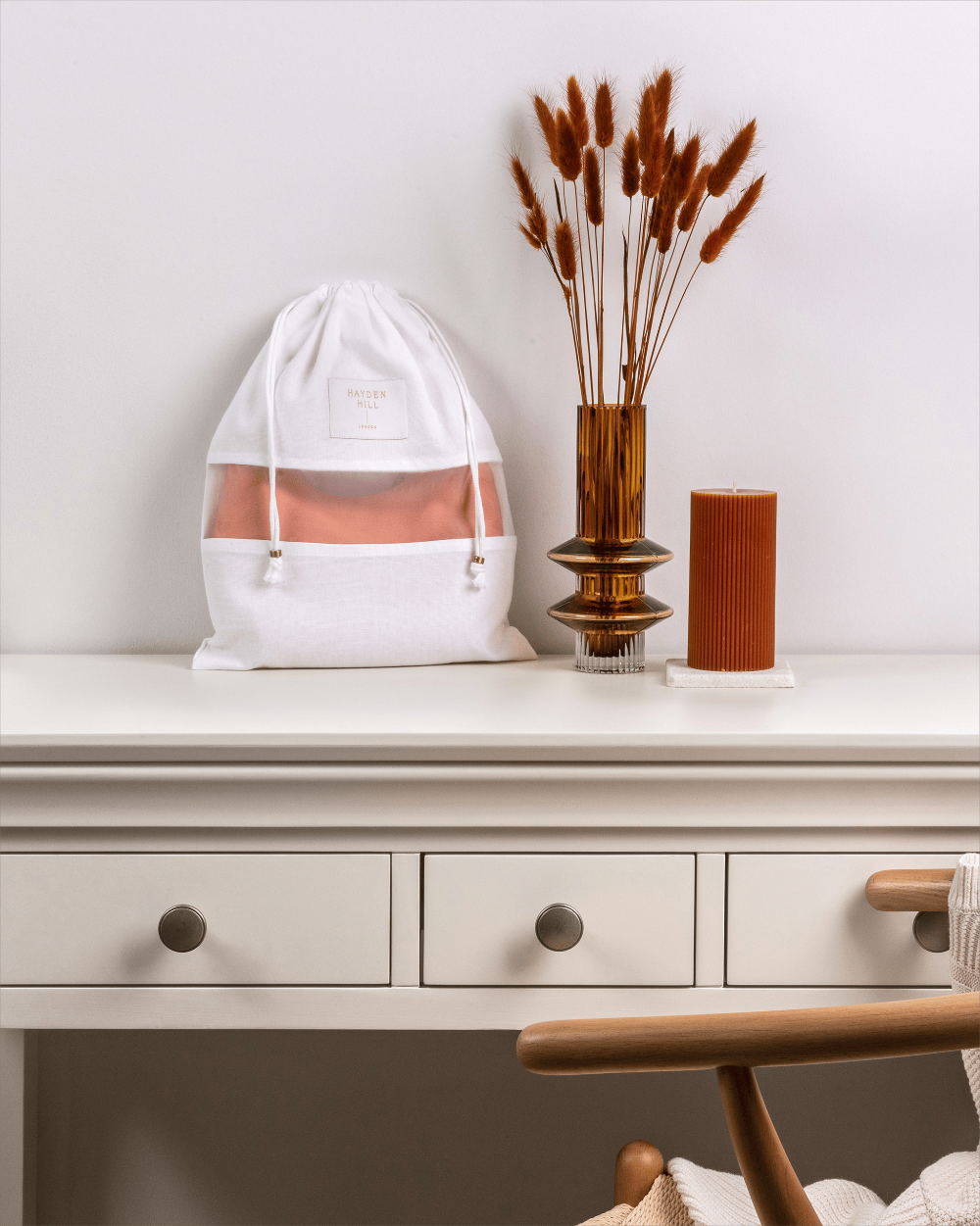 A white drawstring bag with a sheer front window sits on a white table, styled with a brown glass vase of dried grass and a brown candle against a light gray wall.