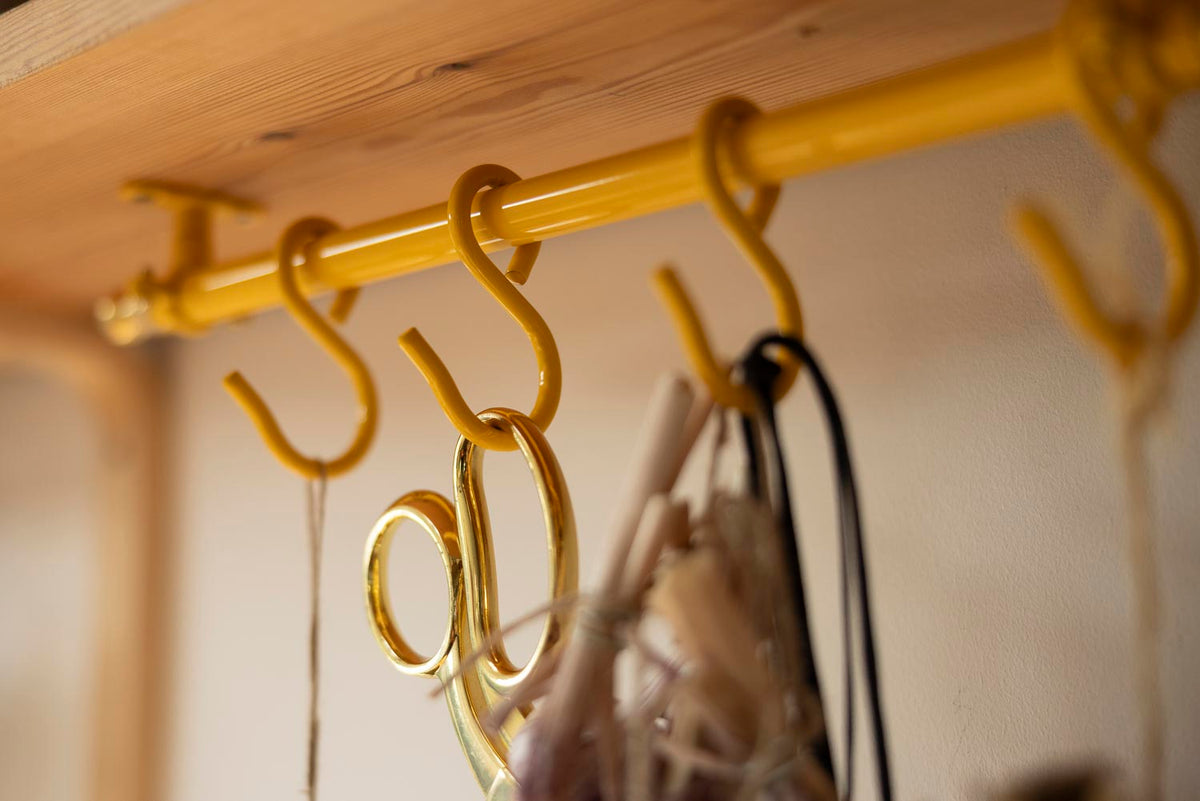 A close-up of a yellow metal rod with multiple hanging hooks mounted under a wooden shelf, with some bags and cords hanging from the hooks.