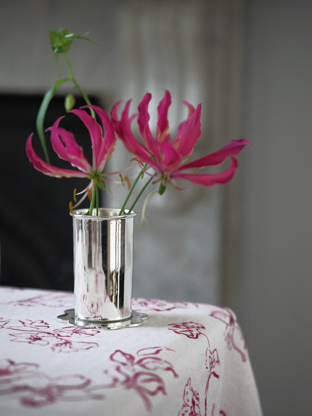 Pink flowers in a silver vase on a table with a floral tablecloth.