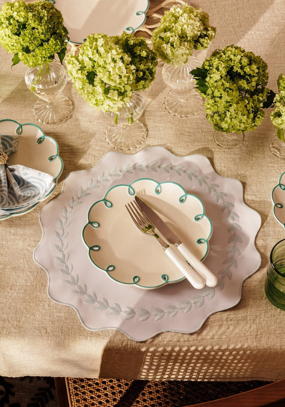 Elegant table setting with decorative plates, silverware, and green floral arrangements on a beige tablecloth.
