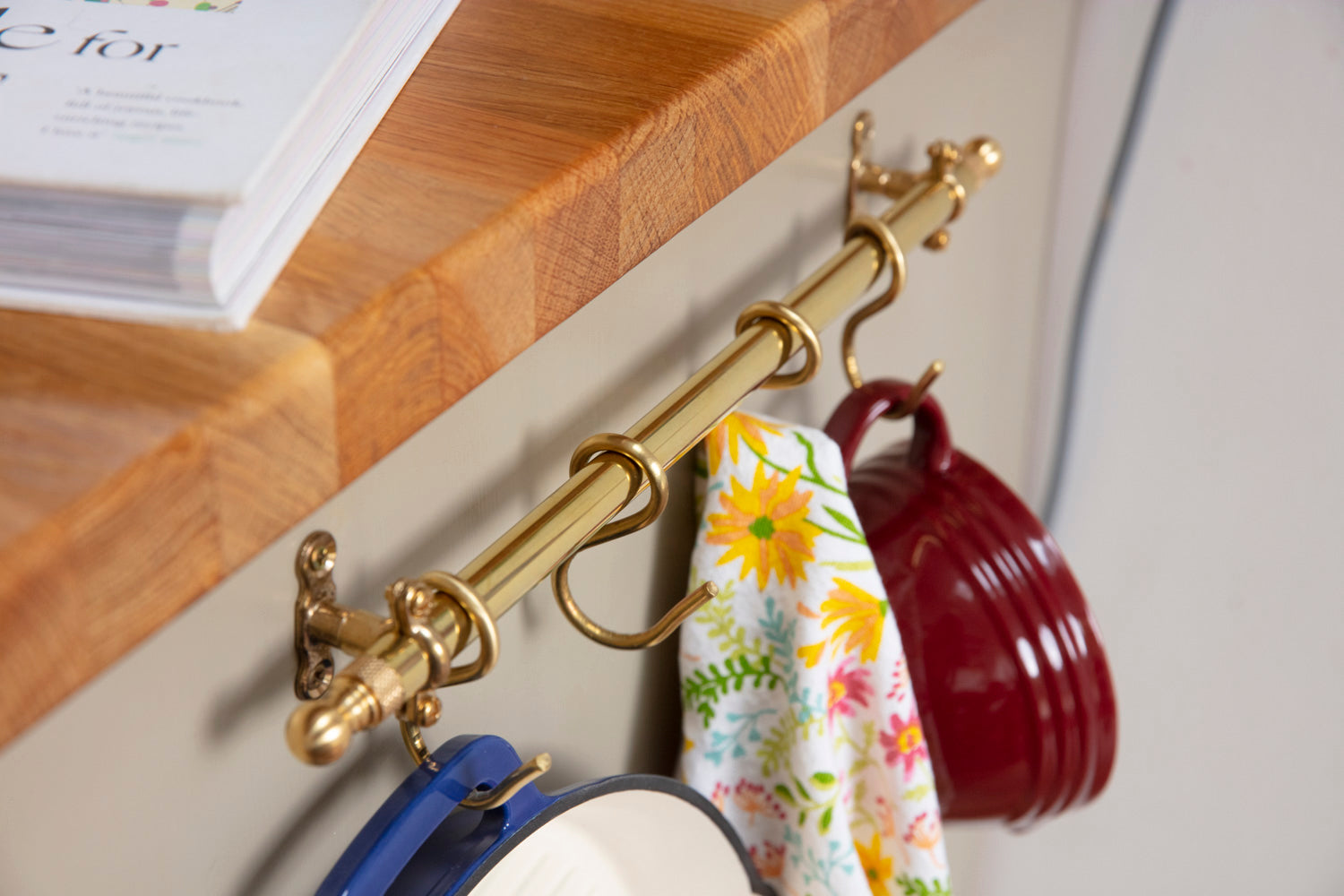 A brass kitchen wall hook rack mounted under a wooden countertop, holding a floral kitchen towel and two hanging mugs, one red and one blue.