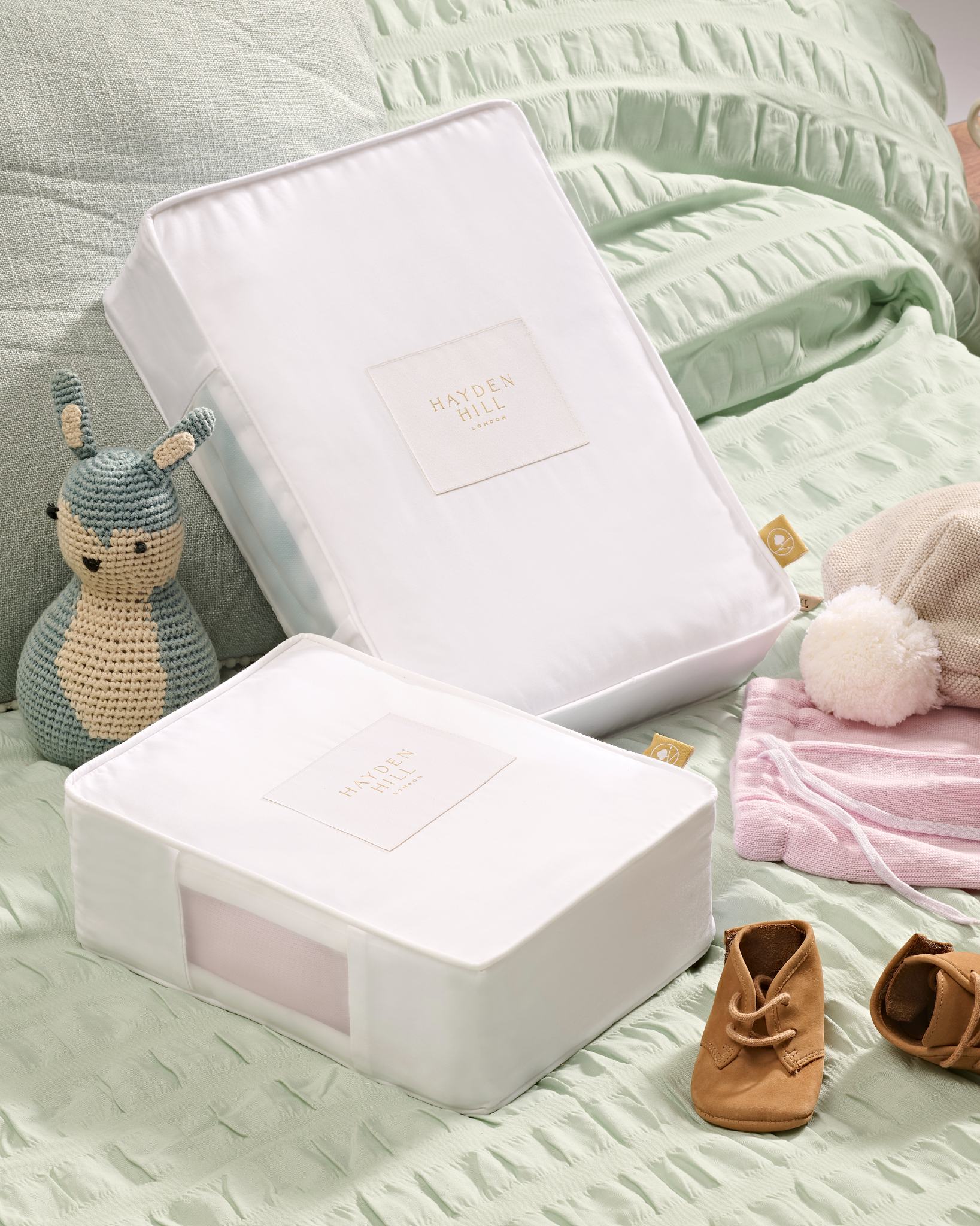 Baby gift set with white boxes, soft toys, and baby shoes on a light green blanket.