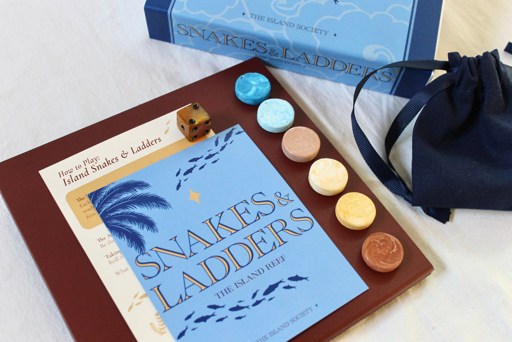 Six coloured Jesmonite playing counters displayed beside The Island Reef Snakes and Ladders instructions and blue illustrated booklet.
