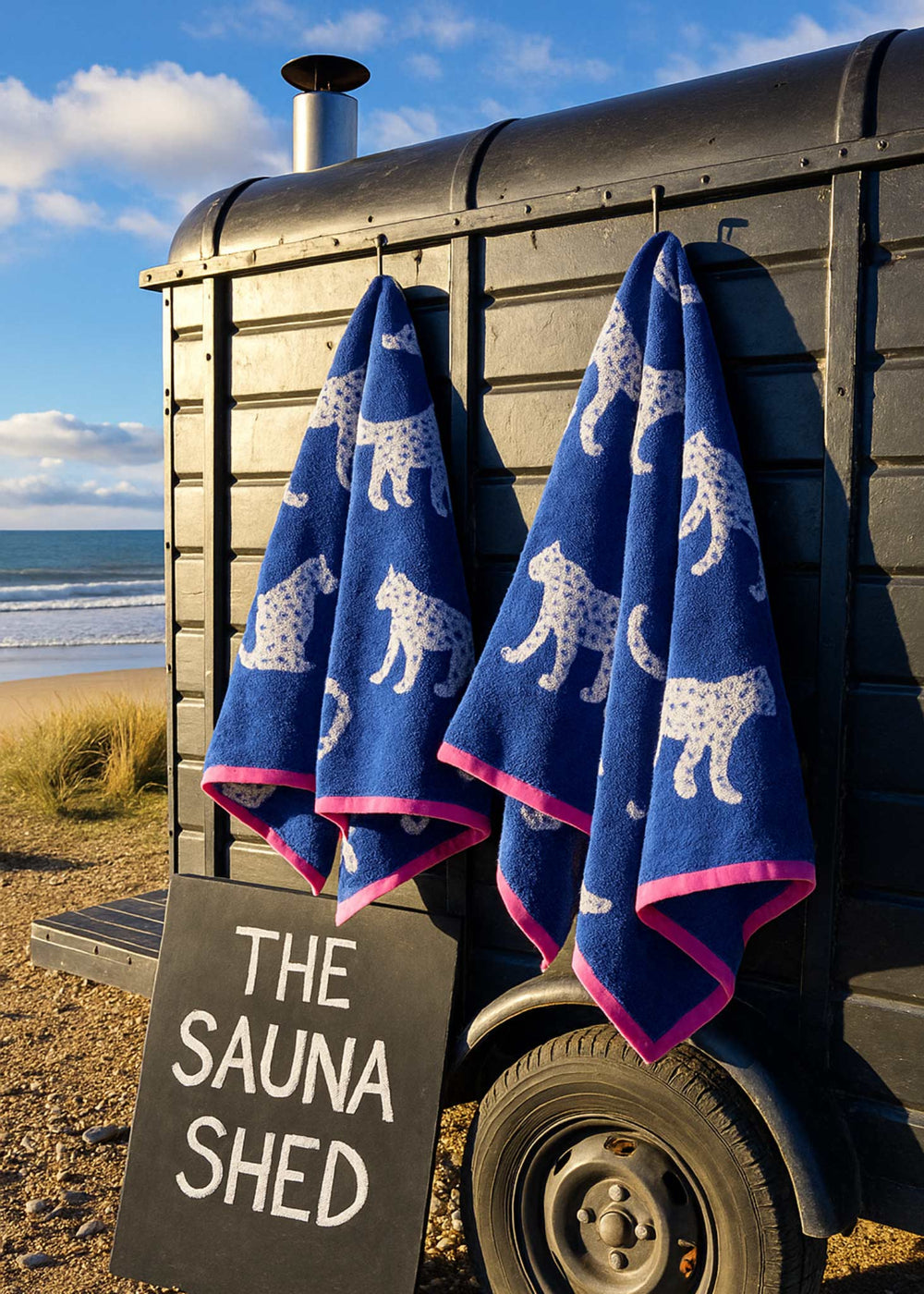 Blue towels with white animal patterns hanging on a trailer labeled 'The Sauna Shed' by the sea.