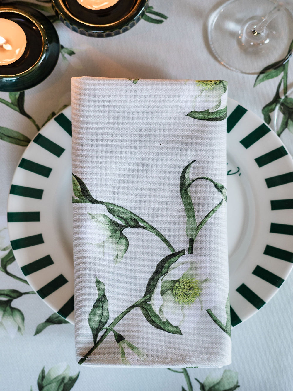 Napkin with floral design on a plate with striped pattern, candles, and glass in the background.