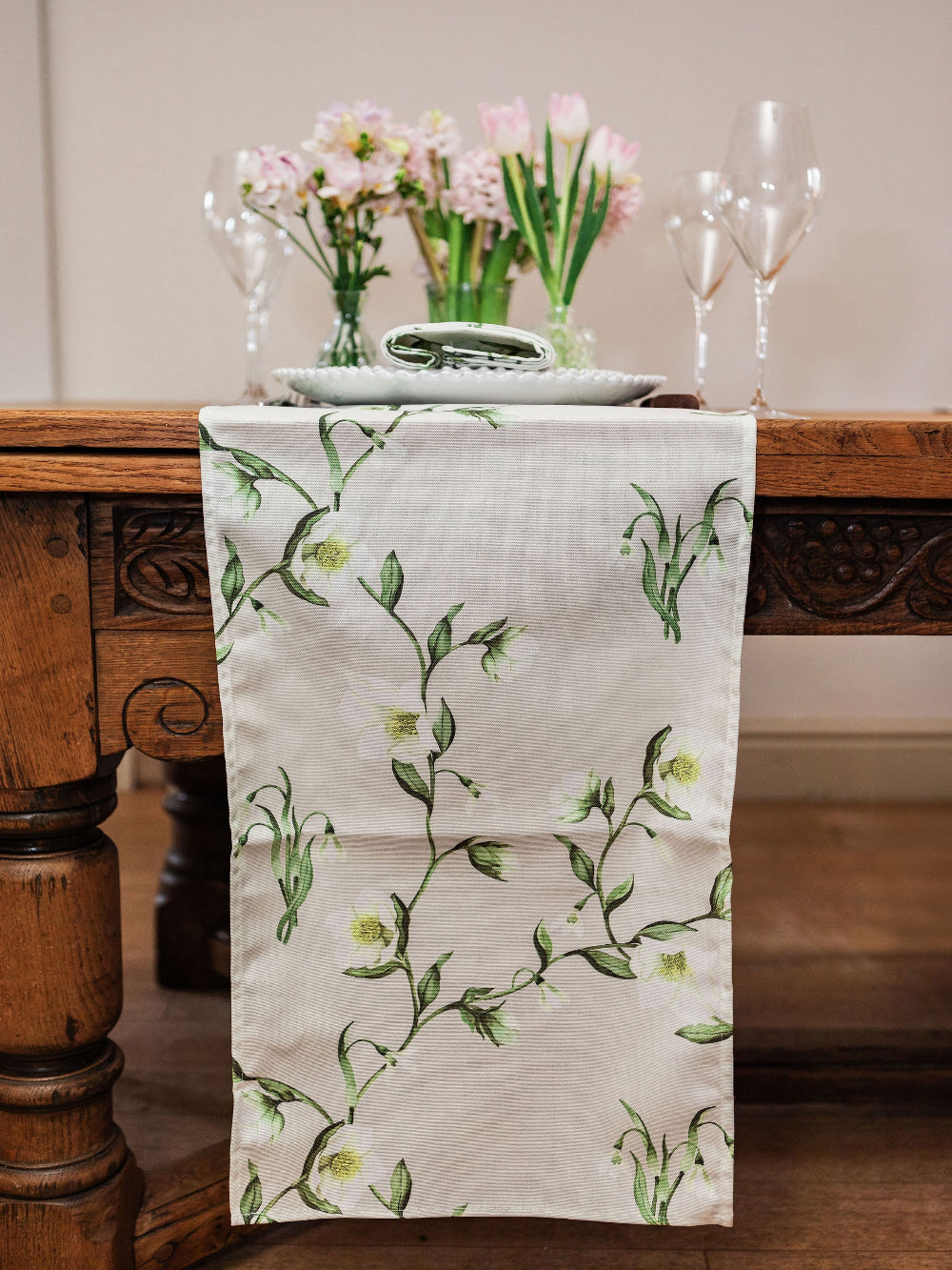 Floral-patterned table runner on a wooden table with flowers and glasses in the background.