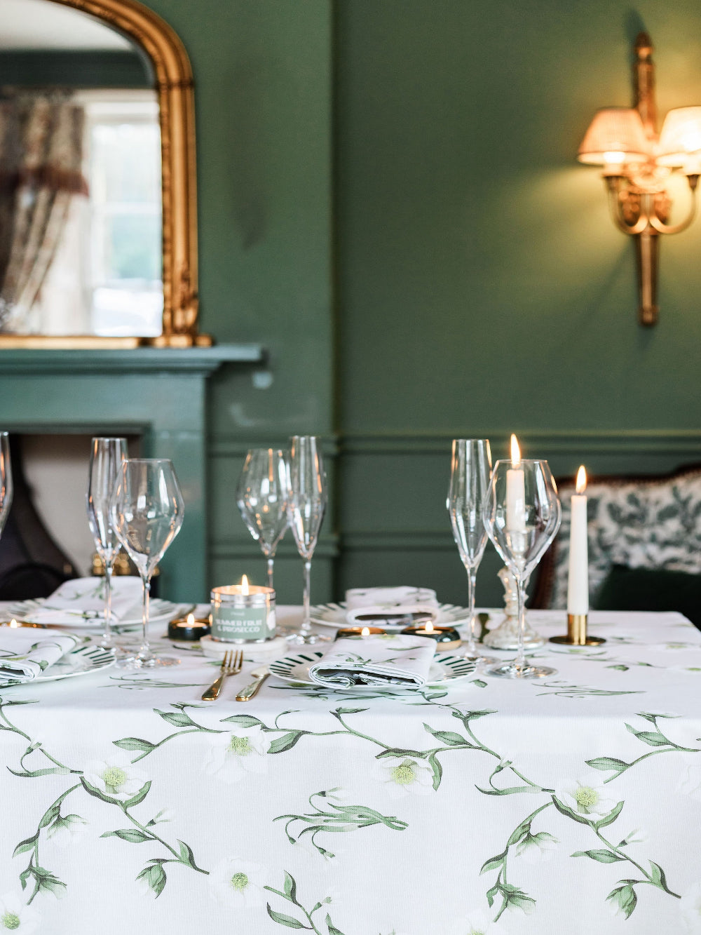Dining table set with glasses, candles, and a floral tablecloth in a room with green walls and a mirror.