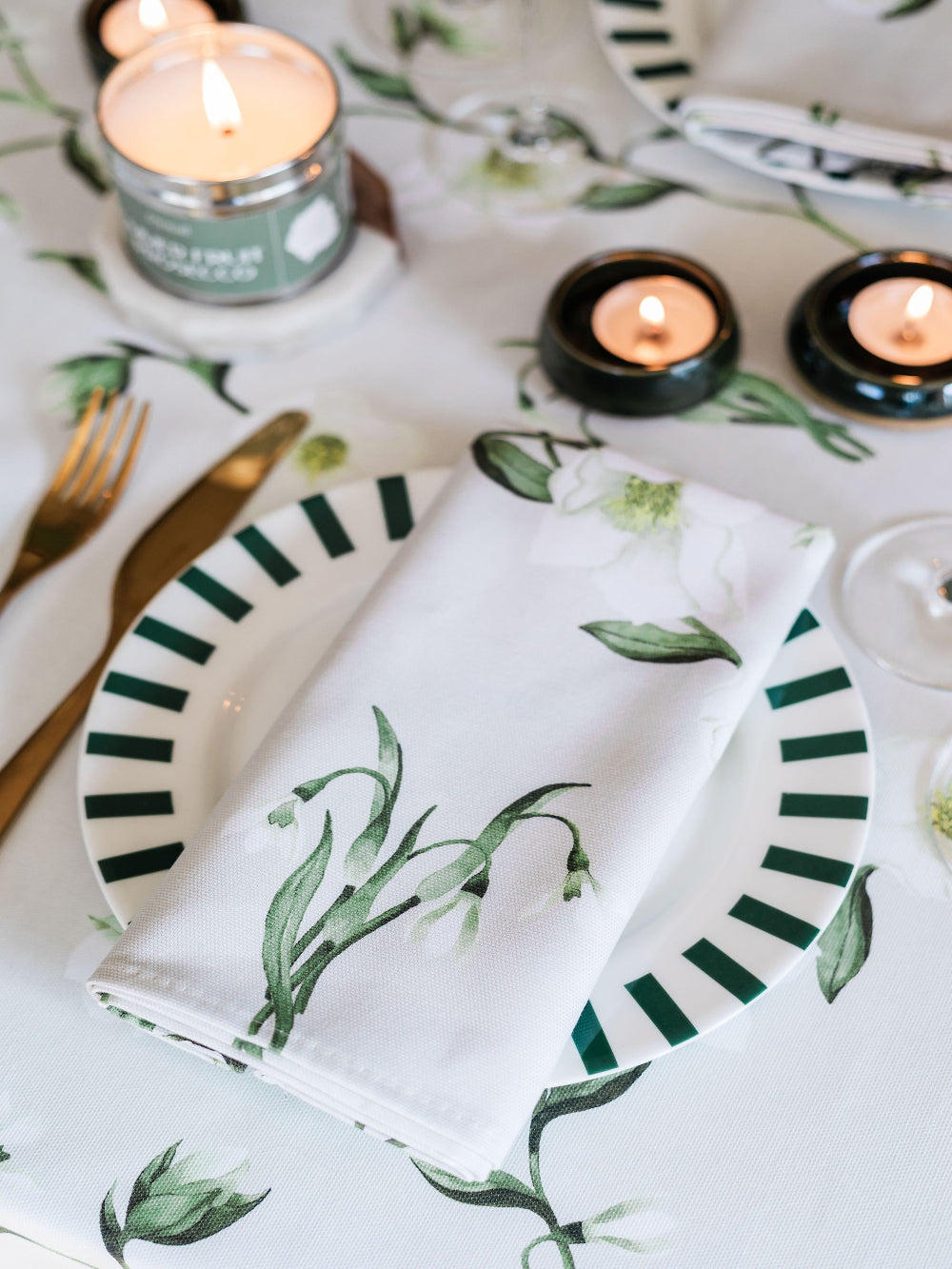 Table setting with green and white patterned plates, napkins, and candles on a floral tablecloth.