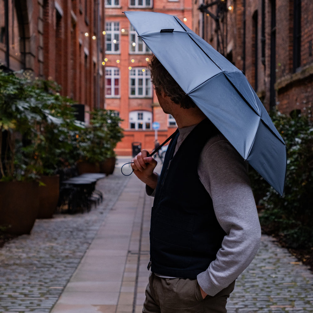 Person holding a blue umbrella in an urban setting with brick buildings and plants.