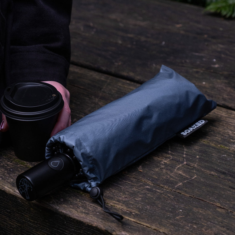 Folded umbrella with a brand logo on a wooden surface, held by a person with a black cup.
