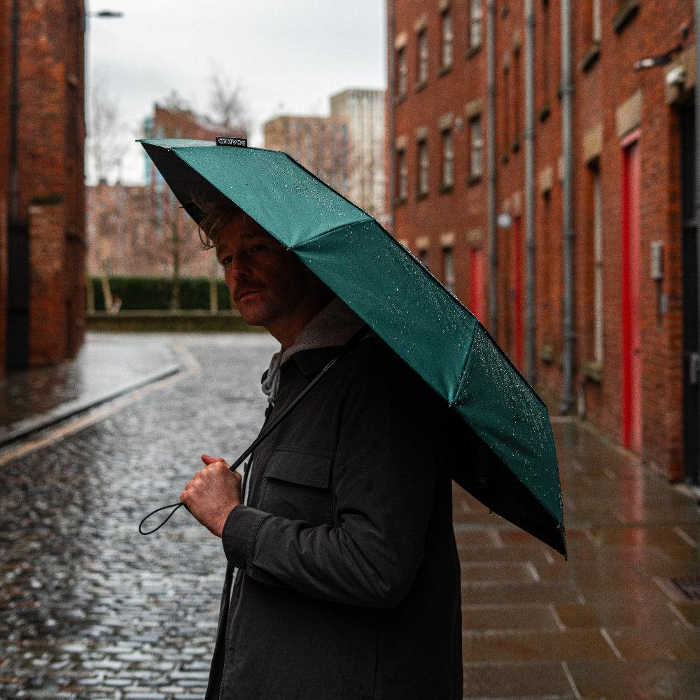 Person holding a green umbrella in a rain-soaked street with brick buildings.