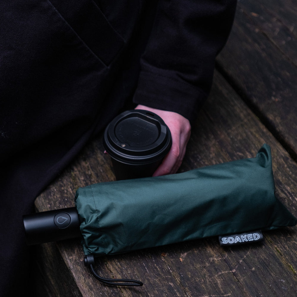 Person holding a black coffee cup with a green folded umbrella on a wooden surface