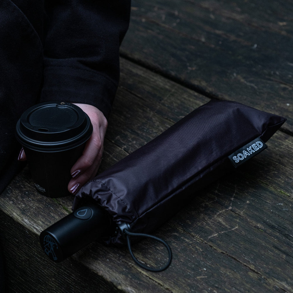 Person holding a black coffee cup with a black umbrella on a wooden surface