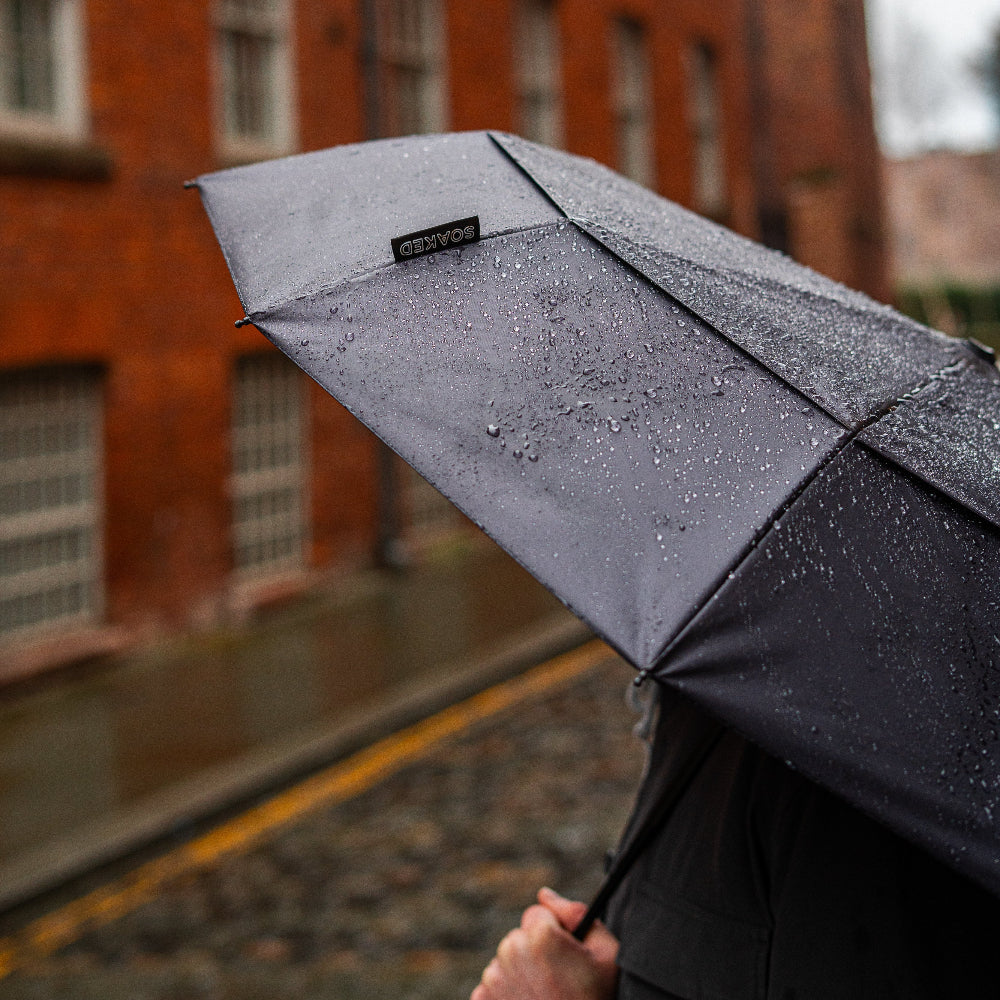Person holding a black umbrella with a brand logo in front of a brick building.