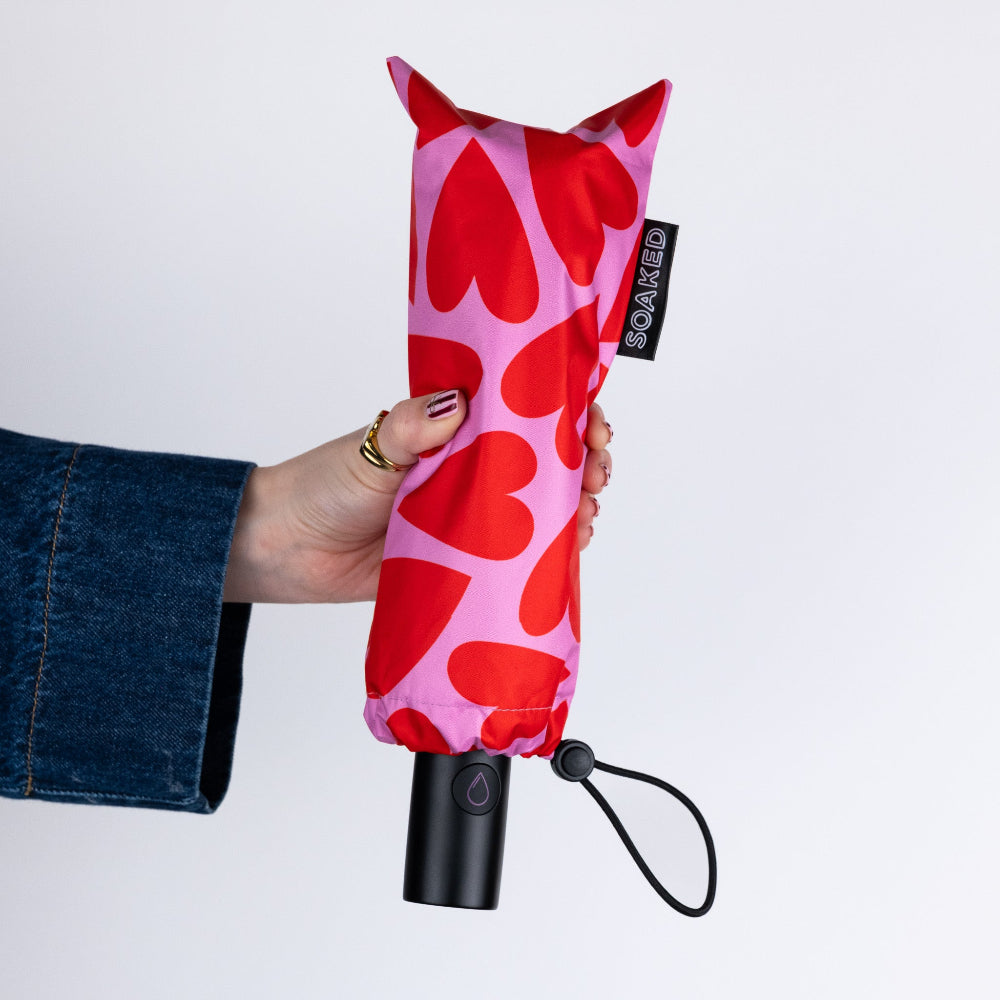 Hand holding a red and pink heart-patterned umbrella with a black handle on a light gray background.