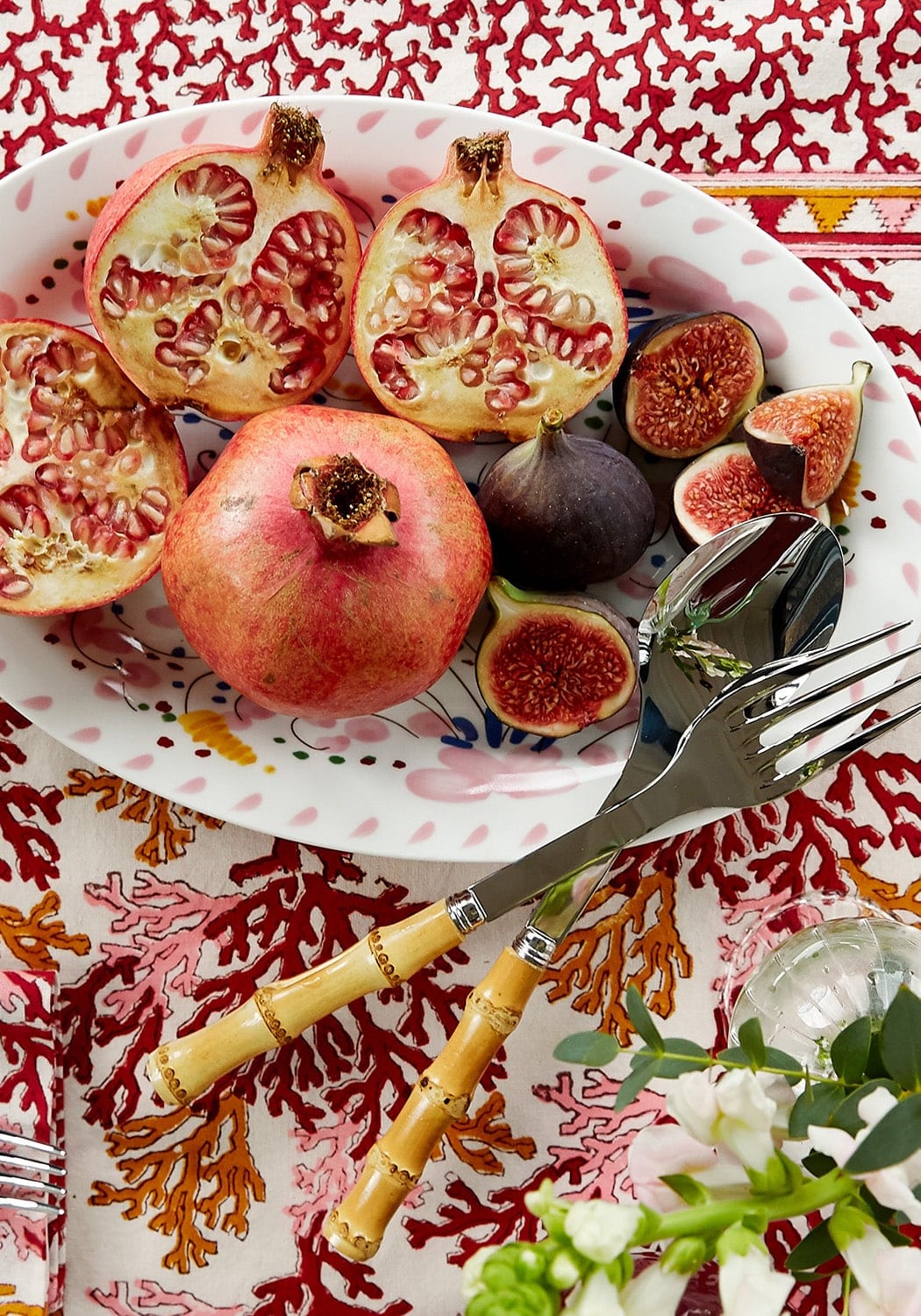 Pomegranate and figs on a decorative plate with cutlery on a patterned tablecloth.