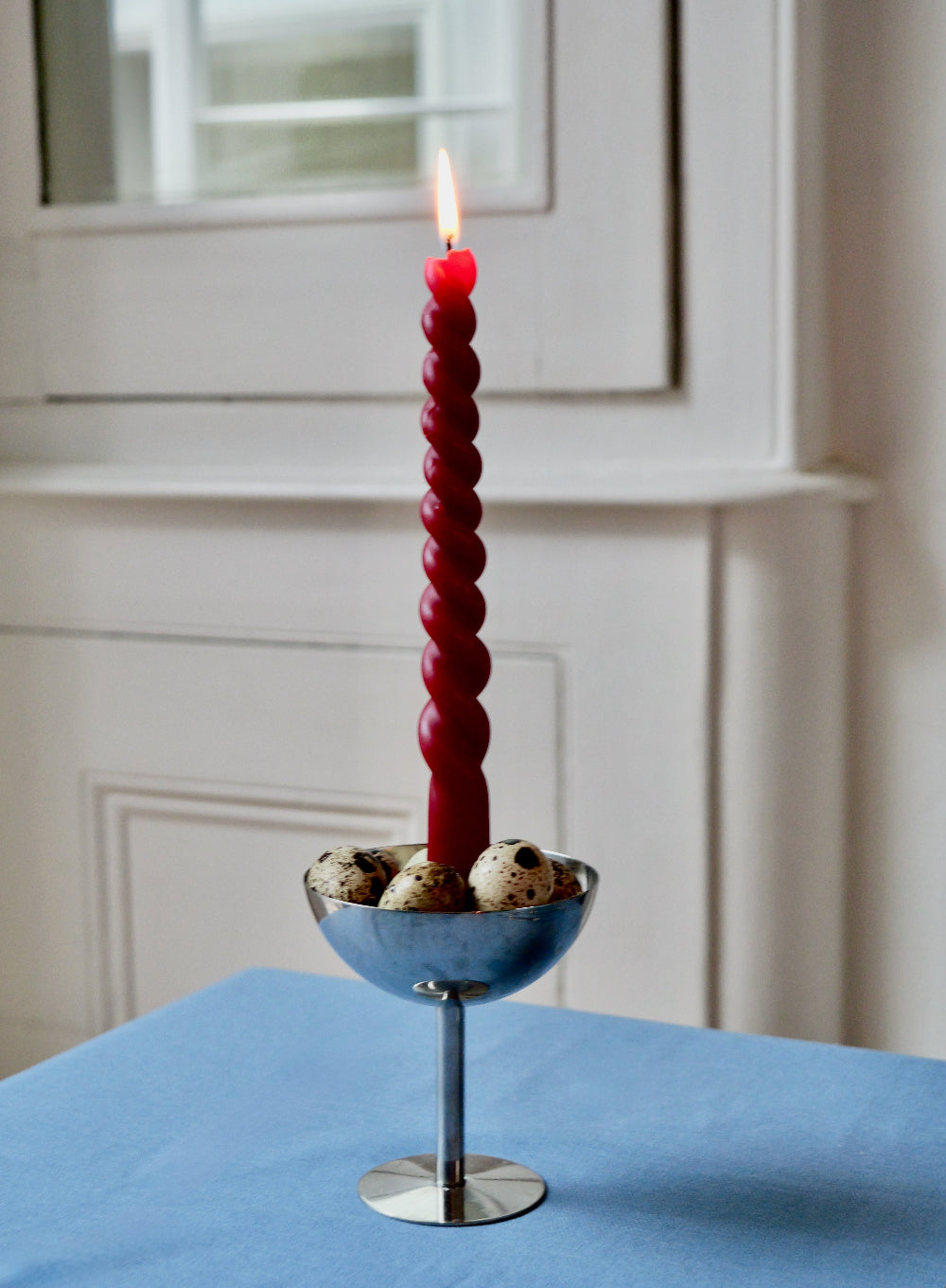 Red twisted candle in a metal desert bowl on a blue surface with a neutral background