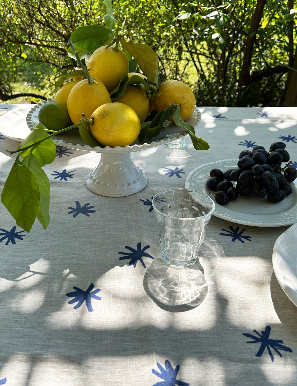 Table setting with lemons on a white pedestal, grapes on a plate, and a clear glass vase on a tablecloth with blue star patterns.