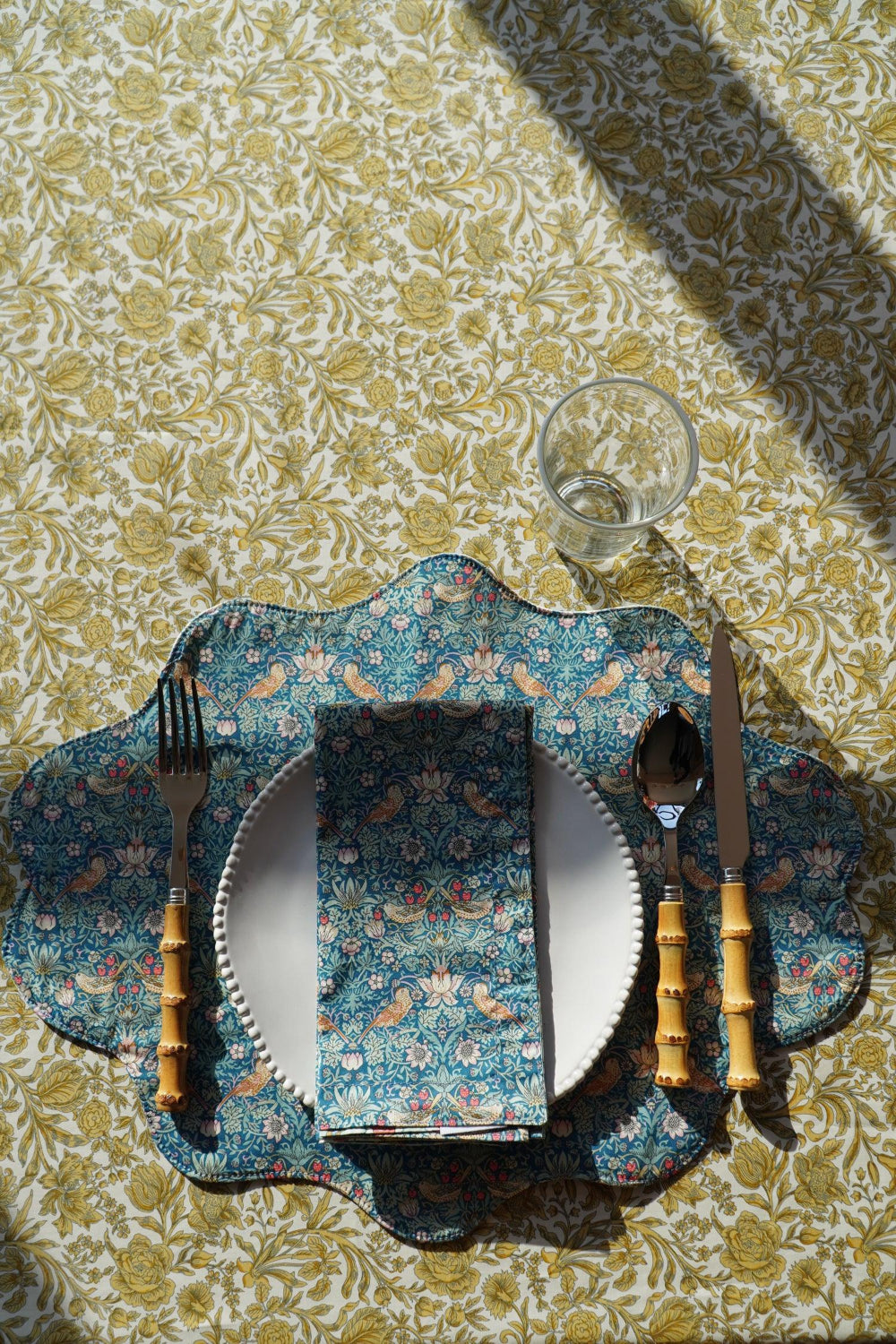 Table setting with floral tablecloth, plate, cutlery, and glass on a patterned surface.