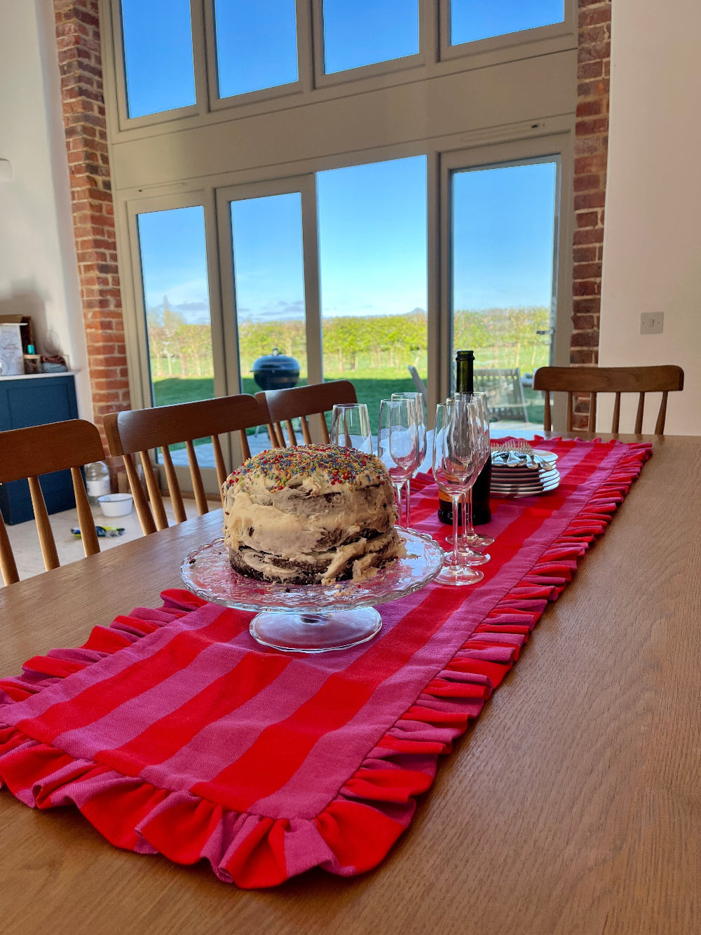 A red and white checkered table runner with ruffled edges is styled on a wooden dining table, featuring a chocolate cake on a glass pedestal, wine glasses, and plates, set in a bright room with large windows and brick pillars.