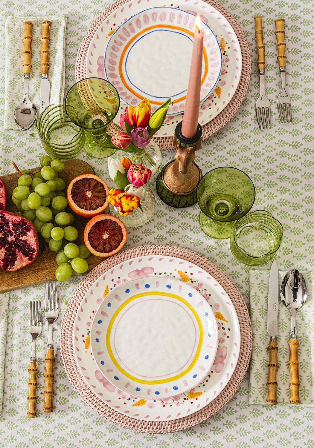 Decorative table setting with colourful plates, glasses, and fruits on a patterned tablecloth.