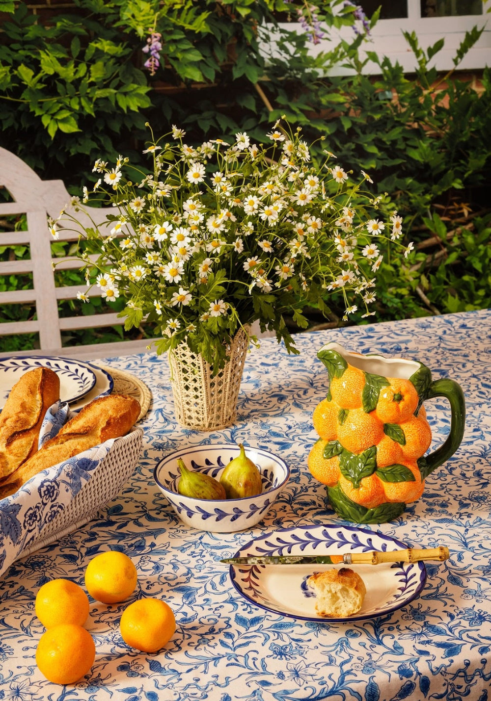 Outdoor table setting with flowers, fruit, and bread on a patterned tablecloth.
