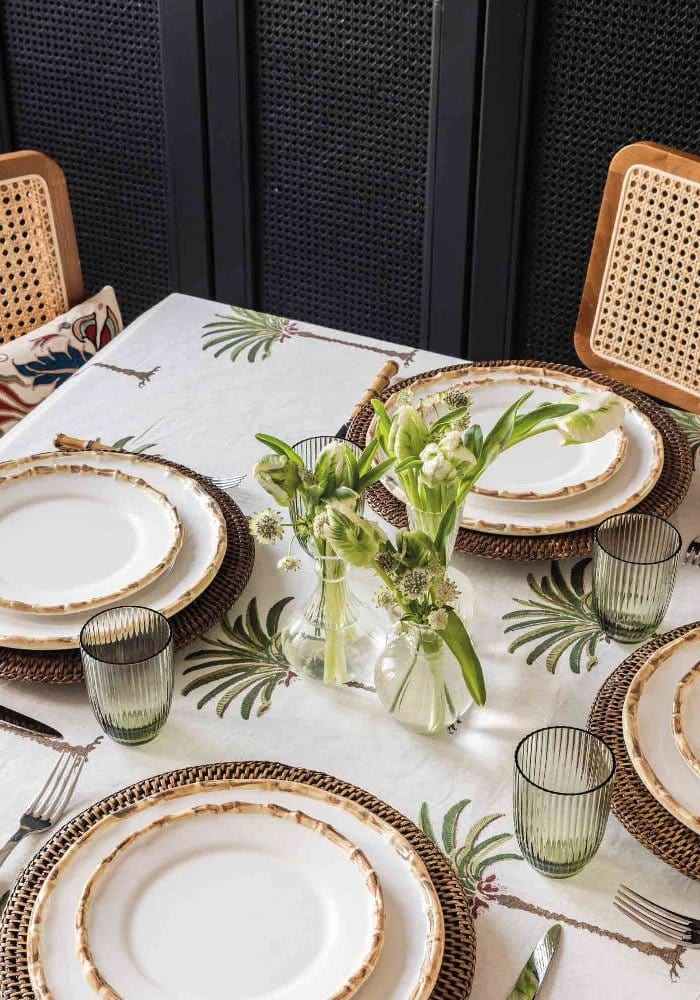 Dining table setting with plates, glasses, and a floral centrepiece on a patterned palm tree tablecloth.
