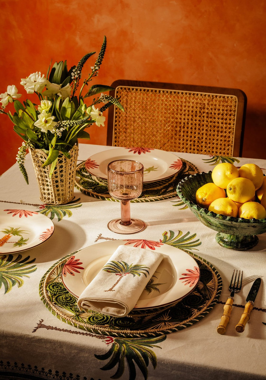 Dining table setting with floral arrangement, lemons, and decorative plates on a patterned palm tree tablecloth.