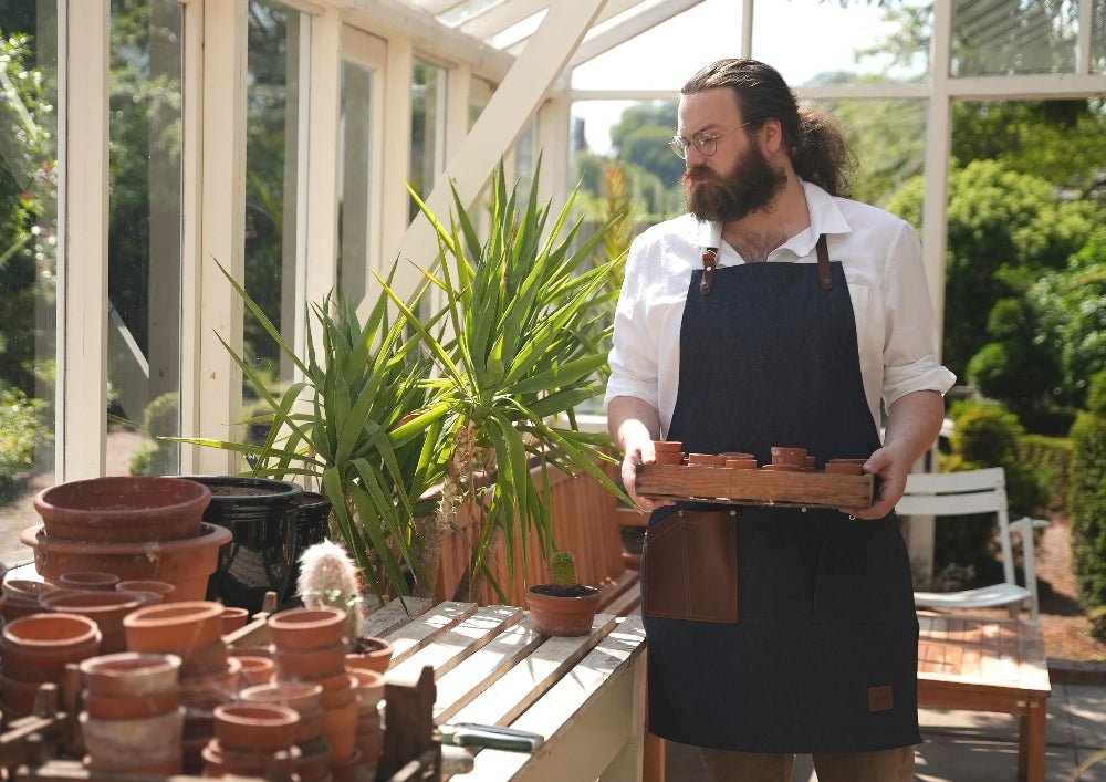 Man holding a tray of terracotta pots in a greenhouse setting with an artisan apron