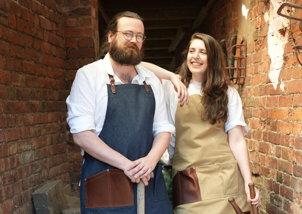 Two people wearing aprons standing against a brick wall.