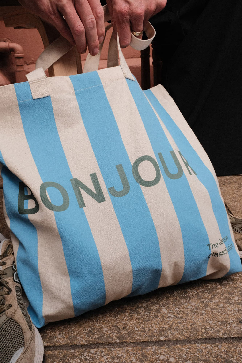 Striped blue and cream tote bag with 'Bonjour' text held by a person on a wooden surface