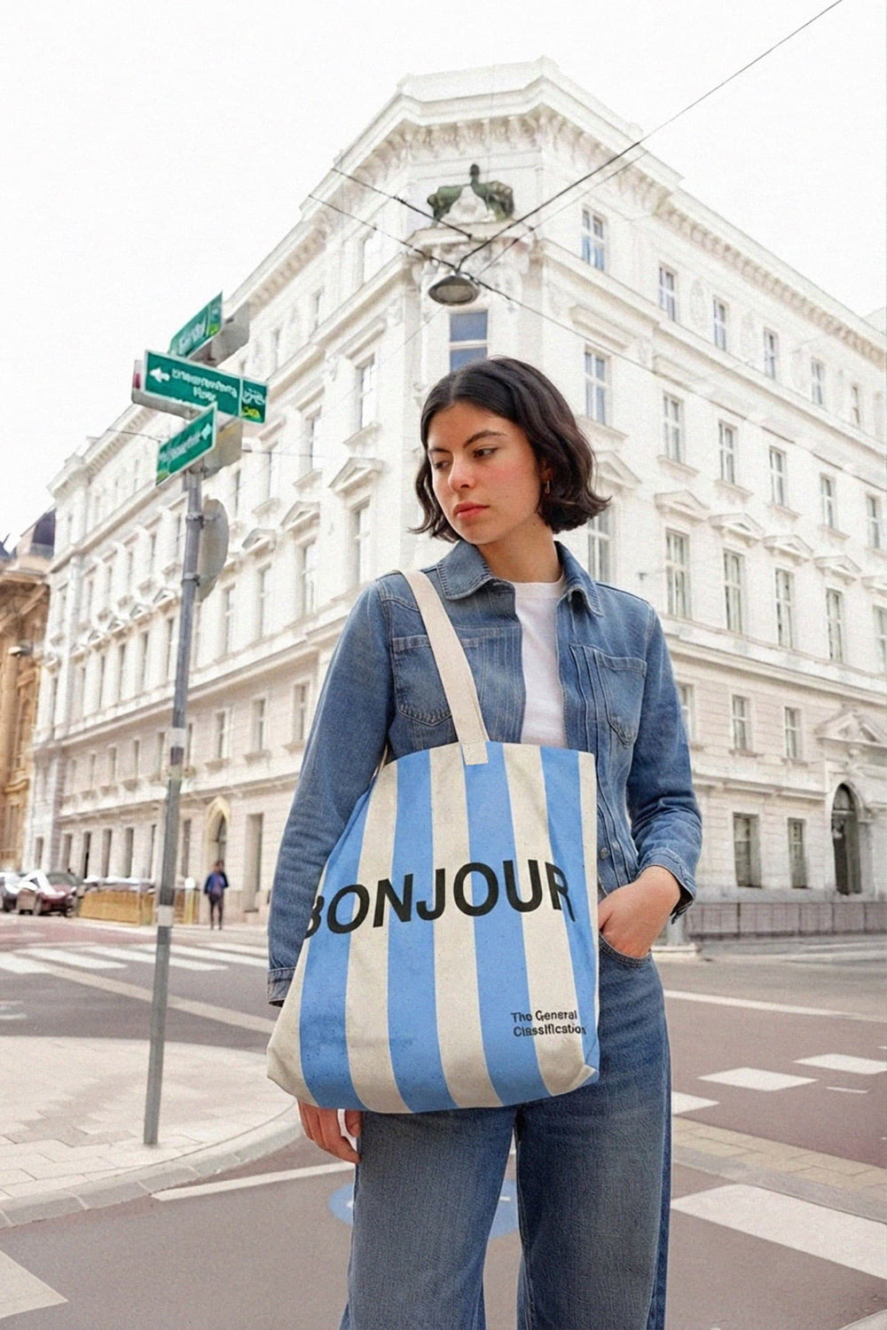 A woman wearing a denim jacket and jeans stands on a paris street holding a blue and white striped tote bag with 'BONJOUR' printed in black. A large white building is in the background.
